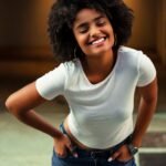 African American woman with afro hair smiling in casual white shirt and jeans, standing outdoors.